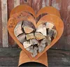 A heart-shaped metal log holder filled with firewood, set against a wooden background. Produktbild-Vorschau
