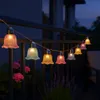 String lights with bell-shaped bulbs in various pastel colors are strung along a balcony railing at dusk. Pink flowers and purple lavender are visible below. Produktbild-Vorschau