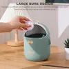 A hand is shown throwing crumpled paper into a small, green trash can with a duck design on a wooden desk. Produktbild-Vorschau