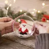 Hands holding a clear Christmas ornament with a small Santa Claus figure inside, surrounded by white decorative snow. Produktbild-Vorschau