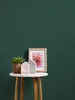 A small round table with a plant, two decorative house-shaped objects, and a framed picture of a flower against a dark green wall. Produktbild-Vorschau