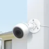 A white security camera is mounted on a white wall, with a blurred house and sky in the background. Voir les images du produit