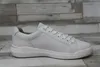 A white leather sneaker is shown from the side against a grey and white wood-paneled background. Náhľad obrázku produktu
