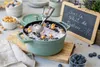 A green Staub pot filled with blueberry ice cream, topped with blueberries and popcorn, sits on a wooden board. A small chalkboard with French writing is in the background. Produktbild-Vorschau