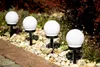 Four white globe-shaped solar lights are placed in a garden bed next to a brick pathway. Produktbild-Vorschau
