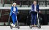 Two people riding electric scooters in front of a modern building. Produktbild-Vorschau