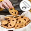 A hand is holding a pretzel-shaped cookie over a basket full of similar cookies. A circular inset shows cookie cutters and a metal ring. Produktbild-Vorschau