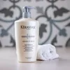 A bottle of Kérastase shampoo and a folded white towel on a countertop, with a decorative tiled wall in the background. Anteprima dell’immagine prodotto