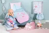 A baby doll is shown with various accessories, including a sleeping bag, pajamas, a potty, and a small plush toy, arranged in a nursery-like setting. Produktbild-Vorschau
