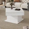 A modern white coffee table with a rotating and lifting top is placed on a textured rug in a living room setting, with a sofa visible in the background. Produktbild-Vorschau
