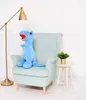A blue dinosaur plush toy sits on a light blue armchair next to a small side table with a potted plant. A gold floor lamp is to the left. Náhled obrázku produktu
