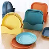 Several stacks of colorful plates and trays are organized in plastic holders on a wooden table. Produktbild-Vorschau
