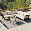 An outdoor patio setting with a sectional sofa, coffee table, and rug, with a stone wall and trees in the background. Anteprima dell’immagine prodotto