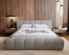 A modern bedroom with a light gray boucle upholstered bed as the main focus. The background features wood paneling and vertical slats, with bedside tables and lighting. Náhled obrázku produktu