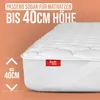 A close-up of a white mattress topper with a red brand tag, set against an orange, wavy background. Text overlays indicate product features. Produktbild-Vorschau