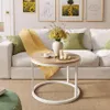 A round coffee table with a wooden top and white metal base, placed in a living room setting with a white sofa, yellow blanket, green pillows, and artwork on the wall. Náhled obrázku produktu
