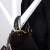 A close-up of a dark brown, patent leather handbag with gold hardware, hanging from a white structure. A dark patterned fabric is visible in the background. Produktbild-Vorschau