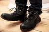 A person wearing black work boots and jeans, standing on a wooden floor. Náhled obrázku produktu
