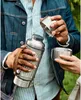 A person holding a silver thermos bottle and a cup in an outdoor setting. Voir les images du produit