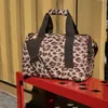 A leopard-print duffel bag sits on a red bench in front of a yellow corrugated metal wall. Náhled obrázku produktu