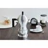 A silver espresso coffee maker is displayed on a wooden table, with a small cup of coffee and a jar of coffee beans in the background. Produktbild-Vorschau