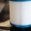 A close-up of a white pleated pool filter cartridge with blue end caps, resting on a dark, reflective surface. Produktbild-Vorschau