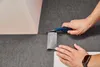Hands are shown cutting a carpet tile with a utility knife, with other carpet tiles and a wall in the background. Produktbild-Vorschau