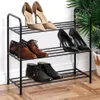 A black metal shoe rack with three shelves, holding various pairs of shoes. The rack is placed on a wooden floor against a white wall, with a large green plant visible in the background. Produktbild-Vorschau