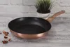 A frying pan with a copper exterior and black interior is placed on a wooden surface, with a small potted plant in the background. Voir les images du produit
