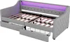 A gray upholstered daybed with wooden slats and two pull-out drawers, one of which is partially open, revealing a wire basket. The headboard has integrated purple LED lighting. Produktbild-Vorschau