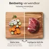 A wooden cutting board is shown divided into two sections. One section displays chopped vegetables (onions, cucumbers, carrots) and herbs, while the other section shows a raw cut of meat. Text overlays describe the features of the board. Produktbild-Vorschau