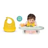 A baby is sitting in a high chair eating broccoli, with a bib and a cup of juice. A yellow bib is shown separately on the left. Produktbild-Vorschau