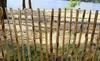 A wooden fence made of natural branches is shown in the foreground, with a body of water and trees in the background. Produktbild-Vorschau