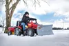 A child is riding a red toy tractor with a snow plow attachment in a snowy field. Produktbild-Vorschau