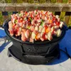 A cast iron grill pot filled with raw meat and vegetable skewers, set up outdoors on a table with a wooden fence in the background. Podgląd zdjęcia produktu
