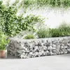 A gabion wall filled with stones, with plants growing around and on it, set against a white wall. Náhled obrázku produktu