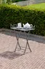 A small metal table with a teapot and cup on it, set on a brick patio with a hedge in the background. Produktbild-Vorschau