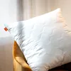 A white quilted pillow rests on a brown chair, with sheer curtains in the background. Produktbild-Vorschau