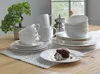 A set of white ceramic dishes, including cups, saucers, bowls, and plates, are arranged on a wooden table with a white lace placemat. A slice of chocolate cake with frosting is on one of the plates, and a small decorative tree is in the background. Produktbild-Vorschau