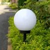 A white spherical garden lamp on a black pole, surrounded by green bushes, with a paved path in the background. Voir les images du produit