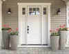 A white front door with glass panes, flanked by two large gray planters with pink flowers, on a gray siding house. Produktbild-Vorschau