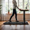 A woman is walking on a treadmill while working at a standing desk in a bright room with large windows. Produktbild-Vorschau