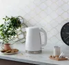 A white electric kettle with a honeycomb texture is placed on a marble countertop next to a cup of tea and cookies, with a clock and a plant in the background. Produktbild-Vorschau