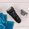 A black electric shaver with blue accents, wet with water droplets, lies on a light gray wooden surface next to two folded blue towels. A protective cap for the shaver is placed to the right. Anteprima dell’immagine prodotto