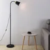 A black and gold floor lamp with a flexible neck stands next to a small round side table with a cup and saucer on it, in a room with a couch visible on the right. Náhľad obrázku produktu
