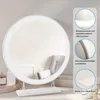 A round makeup mirror with LED lighting on a white stand, placed on a surface with decorative items. Voir les images du produit