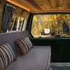 Interior view of a camper van with a sofa and pillows, looking out a window at a campsite with trees and another camper. Anteprima dell’immagine prodotto