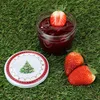 A jar of strawberry jam with a strawberry on top, two whole strawberries, and a lid with a Christmas tree design, all placed on green grass. Produktbild-Vorschau