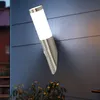 A modern outdoor wall lamp with a motion sensor mounted on a dark gray, horizontally-paneled wall. Some green foliage is visible at the bottom. Produktbild-Vorschau