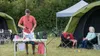 A family camping scene with a man preparing food at a table, while a woman and child relax in chairs near a tent. Anteprima dell’immagine prodotto